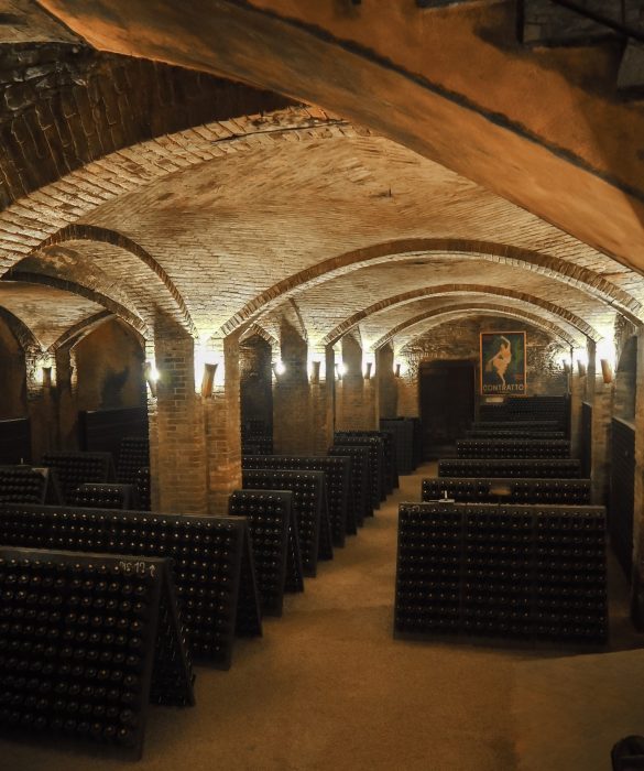 "Interno della Cantina Contratto  con volte a botta in tufo, illuminate da luci soffuse, e bottiglie di spumante metodo classico in affinamento”
