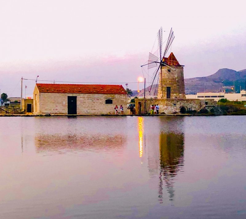"Saline di Trapani al tramonto con vasche d'acqua rosa e arancione, mulini a vento storici in controluce per un weekend romantico in Sicilia"