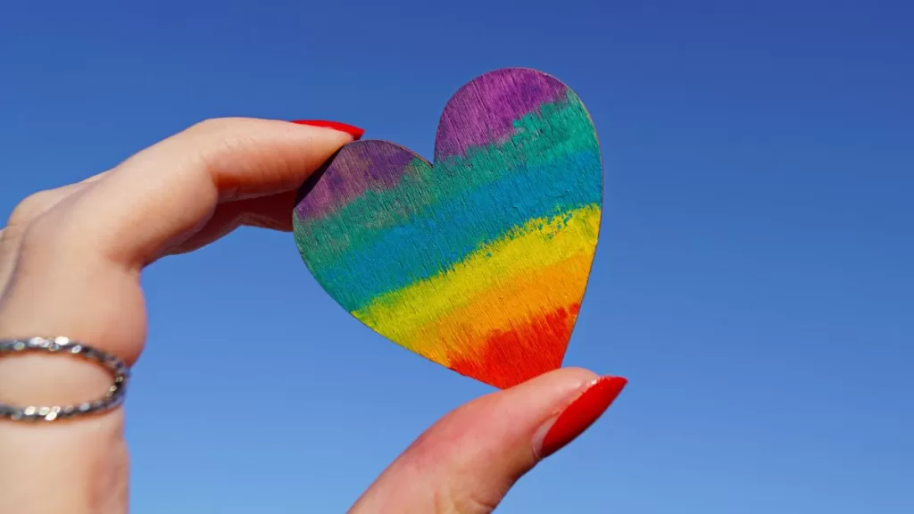 "Mano con unghie rosse che tiene un cuore arcobaleno davanti a un cielo azzurro, simbolo di amore e orgoglio nelle destinazioni LGBTQIA+"